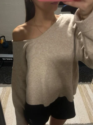 Beige oversized stickad tröja - Mysig beige stickad tröja med avslappnad passform och bred v-ringning som kan bäras off-shoulder. Tröjan har långa ärmar och ribbade muddar vid ärmslut och nederkant. Perfekt för lager på lager och chill dagar.