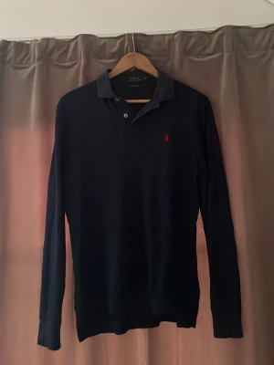 Marinblå långärmad pikétröja Polo Ralph Lauren - Klassisk marinblå långärmad pikétröja från Polo Ralph Lauren med röd broderad logga på bröstet. Tröjan har krage, knappar vid halsen och är tillverkad i mjukt material. Perfekt för en stilren och avslappnad look. Pris går att diskutera😃