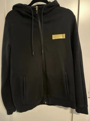 Svart hoodie från HUGO BOSS med dragkedja - Säljer en svart hoodie från BOSS med dragkedja framtill och justerbar huva med snören. På bröstet finns en guldfärgad BOSS-logga. Hoodien har två fickor fram och är tillverkad i mjukt material, perfekt för en avslappnad stil.
