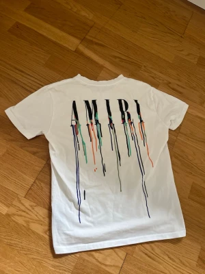 AMIRI Tröja - Säljer en vit t-shirt från AMIRI med färgstänk och tryck både fram och bak. T-shirten har korta ärmar, rund hals och coolt tryck med droppande färger i blått, grönt, orange och svart. Tillverkad i mjuk bomull för skön känsla och stilren look.
