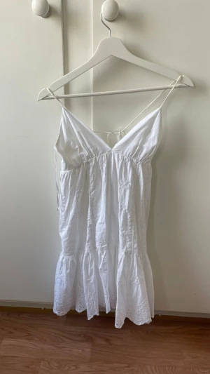 Vit somrig klänning med broderi - Söt vit klänning med tunna axelband och v-ringning. Klänningen har broderade detaljer och volangkant nedtill. Perfekt för varma dagar och har en luftig, avslappnad passform.