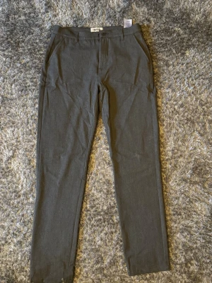 Mörkgrå kostymbyxor från Solid - Snygga mörkgrå kostymbyxor från Solid med smal passform och klassisk design. Byxorna har två sidofickor och bälteshällor i midjan. Perfekta för en stilren look. Materialet känns mjukt och har en lätt stretch för extra komfort.