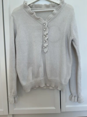 Ljusbeige stickad tröja med volang - Supersöt ljusbeige stickad tröja med volangkant vid halsringning och ärmslut. Tröjan har v-ringning med dekorativa pärlknappar framtill och ribbade muddar. Perfekt för dig som gillar en romantisk och mjuk stil.