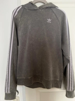 Grå hoodie med ränder från Adidas - Ordinarie pris 850. Har en liten fläck men som säkert går lätt bort i tvätten 🥰Snygg grå hoodie från Adidas med klassiska vita ränder längs ärmarna och Adidas-logga på bröstet. Hoodien har en avslappnad passform, huva och ribbade muddar. Perfekt för en sportig och chill stil.