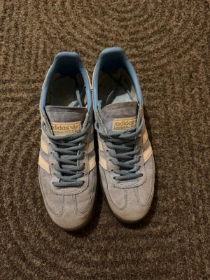 Adidas Handball Spezial blå sneakers - Snygga Adidas Handball Spezial sneakers i ljusblå mocka med klassiska vita ränder på sidorna och mörkbruna gummisulor. Skorna har blå skosnören och en guldfärgad Adidas-logga på plösen. Perfekta för dig som gillar retrostil och streetwear.