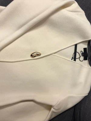 Krämvit kavaj med gulddetalj - Stilren krämvit kavaj med en unik guldfärgad knapp framtill. Kavajen har en minimalistisk design utan synliga fickor och är tillverkad i ett slätt tyg som ger en elegant känsla. Perfekt för dig som gillar en clean och modern look.