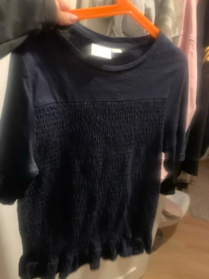 Mörkblå topp med  detaljer - Säljer en mörkblå topp med  detaljer framtill och volangkant nedtill. Toppen har korta ärmar och rund halsringning. Perfekt för dig som gillar en enkel men ändå unik stil. Från Gina tricot