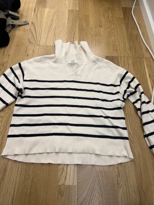 Vit stickad tröja med marinblå ränder - Vit stickad tröja från Gina Tricot med marinblå horisontella ränder. Tröjan har hög ribbad krage som faller ner när man sätter på sig den och långa ärmar. Materialet är mjukt och bekvämt, perfekt för en avslappnad stil. Klassisk och tidlös design som passar till mycket.