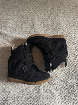Svarta wedge sneakers med kardborre - Säljer ett par svarta sneakers med inbyggd kilklack och kardborreband. Skorna har en cool design med paneler i mocka och textil samt en brun gummisula. Perfekta för dig som vill ha lite extra höjd och streetstyle-vibe. Köparbevis finns!!!