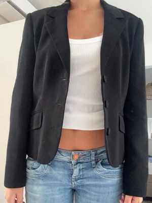 Svart kavaj från DAY Birger et Mikkelsen - Stilren svart kavaj från DAY med klassisk krage och tre knappar framtill. Figurnära passform och två fickor med lock. Perfekt att matcha till jeans eller kjol för en snygg look. Materialet är mjukt och har en lätt stretch.