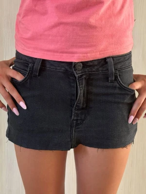 Svart kort jeanskjol med rå kant - Säljer en svart kort jeanskjol i bomull med rå, fransig kant och klassisk femficksdesign. Kjolen har bälteshällor och knappgylf för en avslappnad och trendig vibe.