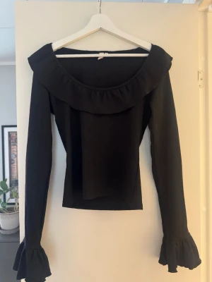 Svart topp med volang och vida ärmar - Säljer en svart långärmad topp med bred volang runt ringningen och vida volangslut vid ärmarna. Toppen har en offshoulder-look och är figurnära i modellen. Perfekt för dig som gillar en trendig och feminin stil.
