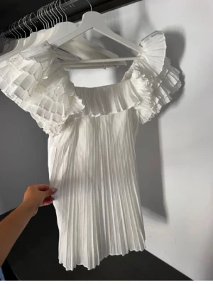 Vit plisserad klänning med volangärm - Supersöt vit klänning med plisserat tyg och stora volangärmar. Klänningen har en rak passform och är tillverkad i ett lätt, luftigt material som ger en elegant look. Perfekt för dig som gillar trendiga detaljer och en romantisk stil.