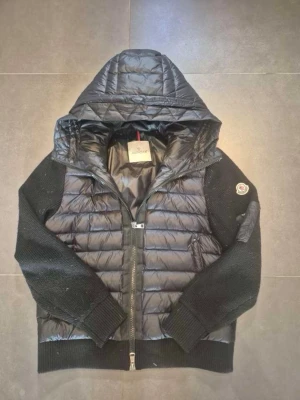 Svart cardiganjacka från Moncler - Snygg svart cardiganjacka från Moncler med quiltad front och huva i glansigt material samt stickade ärmar. Jackan har dragkedja framtill, ribbade muddar och en Moncler-logga på ärmen. Perfekt för dig som vill ha både stil och komfort.size 1 :s