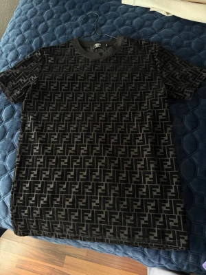 Svart t-shirt med monogrammönster - Snygg svart t-shirt med all-over monogrammönster i mörkgrått. T-shirten har rund halsringning och korta ärmar. Materialet ser ut att vara mjukt och bekvämt, perfekt för en trendig streetwear-look.