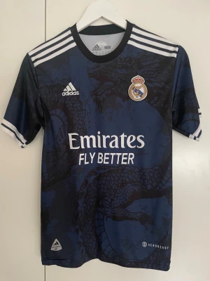 Real Madrid bortatröja Vini Jr 20 - Mörkblå Real Madrid bortatröja med drakmönster, vita detaljer och tryck. På bröstet finns klubbmärket och Adidas-loggan, samt sponsortryck. På ryggen står det VINI JR. och nummer 20. Tillverkad i lätt och ventilerande material.