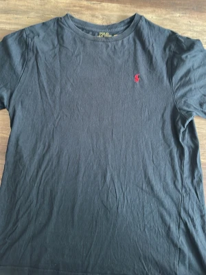 Svart t-shirt från Polo Ralph Lauren - Barnstrolek i XL. Klassisk svart t-shirt från Polo Ralph Lauren med röd broderad logga på bröstet. T-shirten har rund halsringning och korta ärmar. Tillverkad i mjuk bomull som känns skön mot huden. Perfekt till jeans eller shorts.