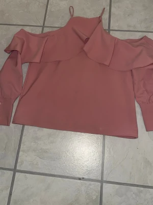 Rosa offshoulder topp med volang - Säljer en ljusrosa offshoulder topp med bred volangdetalj över bröstet och axlarna. Toppen har långa ärmar med resår vid ärmsluten och tunna axelband. Perfekt för dig som gillar en feminin och trendig stil.