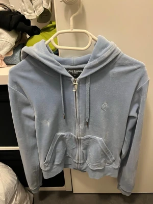 Ljusblå Juicy Couture kofta - Säljer en ljusblå zip-hoodie från Juicy Couture i mjuk velour. Hoodien har huva med dragsko, fickor framtill och en snygg J-dragkedja. Liten Juicy Couture-logga broderad på bröstet. Perfekt för en chill och trendig look.
