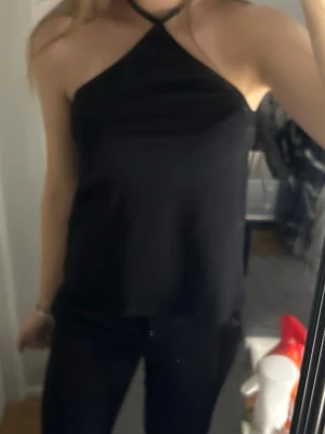 Svart halterneck topp med knyt - Snygg svart halterneck topp med knyt i nacken och öppen rygg. Toppen har en loose passform och är perfekt för dig som gillar minimalistisk stil. Tillverkad i ett mjukt och lätt material som känns skönt mot huden.