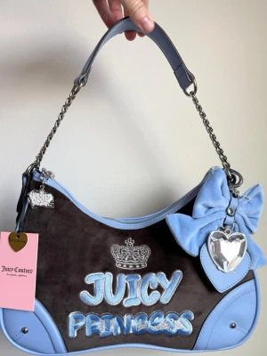 Juicy Couture blå och brun handväska - Snygg handväska från Juicy Couture i brun sammet med ljusblå detaljer i skinnimitation. Väskan har kedjehandtag, stor blå rosett, hjärtformad nyckelring och texten 'JUICY PRINCESS' broderad framtill. Krona i strass och silverfärgade detaljer ger extra bling.