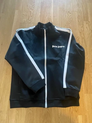 Svart track jacket från Palm Angels - Svart track jacket från Palm Angels med vita ränder längs ärmarna och vit dragkedja. Tröjan har hög krage, ribbade muddar och logga på bröstet. Perfekt för en sportig och trendig look.