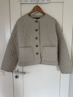 Beige quiltad cardiganjacka från Zara - Säljer en beige quiltad cardiganjacka från Zara med rund halsringning och stora bruna knappar. Jackan har två rejäla framfickor och ett kortare, boxigt snitt. Materialet är mjukt och lätt, perfekt för lager-på-lager under kyliga dagar.