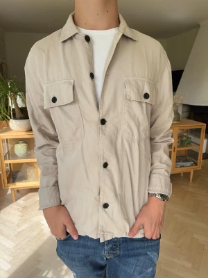 East west overshirt beige creme herr - Snygg beige overshirt från East West! ✨En perfekt skjortjacka/overshirt i en stilren ljusbeige färg. Utrustad med dubbla bröstfickor och snygga kontrasterande mörka knappar. Väldigt mångsidig och lätt att styla över en t-shirt till både vardag och fest. Mycket fint skick!#eastwest #overshirt #skjortjacka #herrkläder #vårjacka #höstjacka #streetwear #casual #beige #skjorta #secondhand