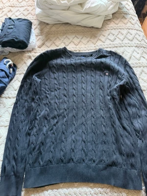 Mörkblå kabelstickad tröja från GANT - Snygg mörkblå kabelstickad tröja från GANT med rund halsringning och diskret logga broderad på bröstet. Tröjan har långa ärmar och ribbade muddar vid ärmslut och nederkant. Perfekt för dig som gillar klassisk och stilren design.