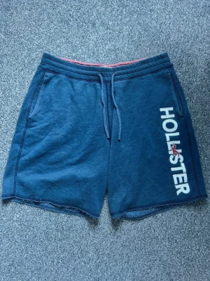 Hollister Shorts  - Blå mjuka shorts från Hollister med vit logga och röd detalj på ena benet. Shortsen har snörning i midjan, två sidofickor och råa benslut. Perfekta för en avslappnad stil under varmare dagar. 