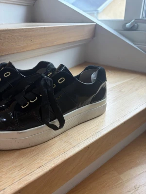 Svarta lackade sneakers från Gant - Snygga svarta sneakers från Gant i lackat material med vita sulor och guldfärgade detaljer vid snörningen. Klassisk låg modell med svarta breda skosnören och diskret logga på sidan. Perfekta för en trendig och stilren look.