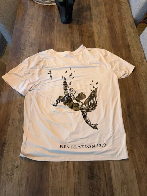 Beige t-shirt med tryck och text - Säljer en beige t-shirt med svart tryck av en fallande ängel och texten 'REVELATION 12:9' på ryggen. Framtill finns ett mindre svart tryck på bröstet. T-shirten har rund hals och korta ärmar, perfekt för dig som gillar unika motiv.