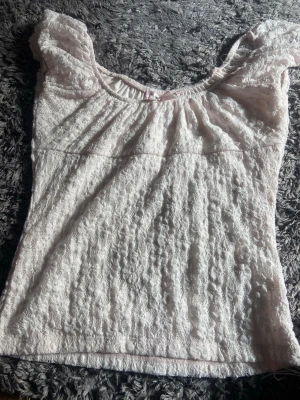 Ljusrosa spetsblus med volang - Supersöt ljusrosa blus med spets och volangdetaljer vid axlarna. Blusen har en liten rosett framtill och är ärmlös med en romantisk känsla. Perfekt för dig som gillar en feminin och söt stil. Ny och i perfekt skick med storleken av 170!