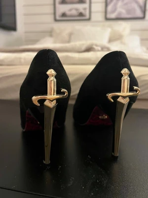 Svarta klackar med gulddetaljer - Unika svarta pumps med stilettklackar i metall och guldfärgade svärdsdetaljer på hälen. Skorna har en elegant spetsig tå och ovandel i sammetsliknande material. Perfekta för dig som vill sticka ut med en edgy och lyxig look.