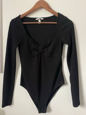 Svart ribbad body med lång ärm - Snygg svart body med lång ärm och djup ringning. Ribbat material och en dekorativ knutdetalj framtill ger en trendig look. Perfekt att styla till jeans eller kjol för en clean outfit.