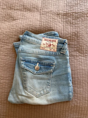 True Religion Billie ljusblå jeans - Säljer ett par ljusblå True Religion Billie jeans med rak passform och mellanhög midja. Klassisk femficksmodell med snygga detaljer på bakfickan och ikonisk patch bak. Perfekta till en avslappnad streetstyle-look.