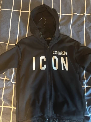 Svart hoodie från Dsquared2 ICON - Svart hoodie från Dsquared2 med dragkedja och stor vit text 'ICON' på framsidan. Är mycket varm och passar Medium och small 