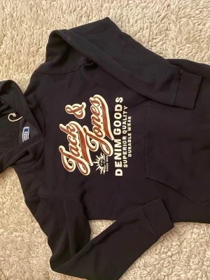 Marinblå hoodie Jack & Jones - Snygg svart hoodie från Jack & Jones med stor vit och beige logga på bröstet där det står 'Denim Goods Superior Quality Durable Wear'. Klassisk känguruficka framtill och huva med dragsko. Perfekt för en avslappnad och trendig stil.