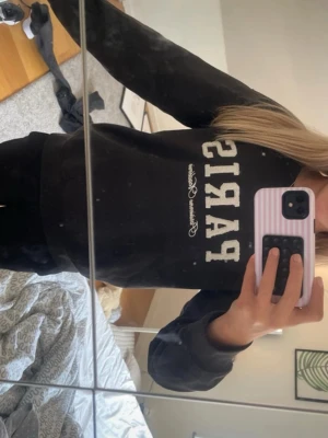 Svart sweatshirt med PARIS-tryck - Säljer en svart sweatshirt med stort vitt PARIS-tryck på bröstet och dekorativ text under. Tröjan har rund halsringning och långa ärmar. Perfekt för dig som gillar enkel men snygg streetstyle. Materialet känns mjukt och bekvämt.