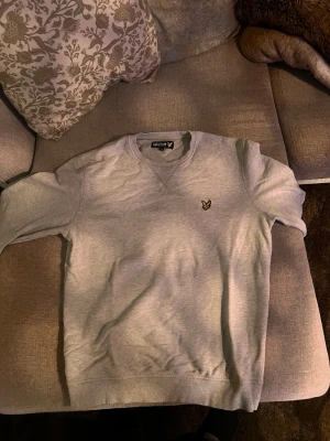 Grå sweatshirt från Lyle & Scott - Snygg grå sweatshirt från Lyle & Scott med klassisk gul logga på bröstet. Tröjan har rund halsringning och ribbade muddar vid ärmslut och nederkant. Perfekt för en avslappnad och stilren look.