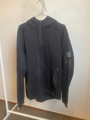 Peak Performance M Tech Zip Hood Herr XL - Säljer en svart hoodie med dragkedja och huva. Modellen har fickor med dragkedja och ett tryck på ena ärmen där det står 'PEAK PERFORMANCE'. Perfekt för en avslappnad och sportig stil.