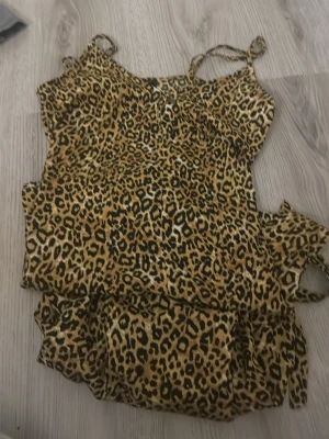Leopardmönstrad lång sommarklänning med en slit - Säljer en leopardmönstrad klänning med tunna axelband och volangdetaljer. Klänningen har en figurnära passform och är gjord i ett lätt, mjukt tyg som känns skönt mot huden. Perfekt för dig som gillar djurmönster och vill sticka ut.