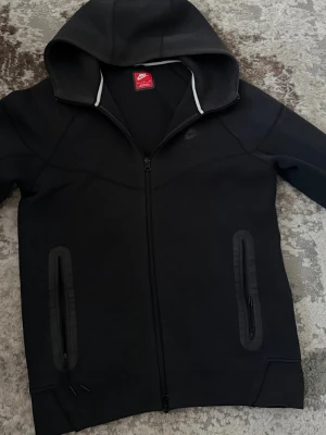 Svart hoodie med dragkedja från Nike - Säljer en svart hoodie från Nike med hel dragkedja framtill och två fickor med dragkedja. Hoodien har en stor huva och diskret Nike-logga på bröstet. Tillverkad i ett mjukt och stretchigt material, perfekt för en sportig och avslappnad stil.