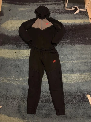 Svart och grå Nike loungewear-set - Säljer ett svart och grått loungewear-set från Nike med huva och dragkedja på tröjan samt matchande byxor med resår i midjan. Båda delarna har Nike-logga i rött. Perfekt för chill dagar hemma eller när du vill vara bekväm. Den är lite solbränd men funkar fortfarande att ha på sig, funkar perfekt för vintern. 