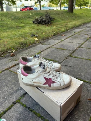 Fina golden goose - Snygga vita sneakers med en rosa glittrig stjärna på sidan och matchande rosa detalj på hälen. Skorna har vita snören, låg profil och texten 'GOLDIGSTAR' på sidan. Tillverkade i läder med slitstark gummisula. Perfekta för en trendig streetstyle-look.