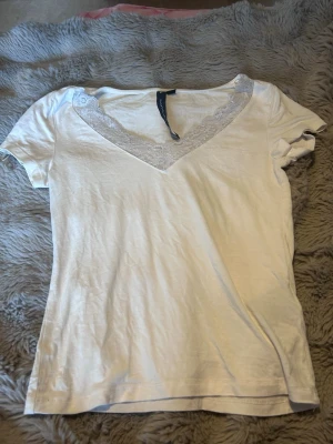 Vit topp med spetsdetalj - Snygg vit topp med v-ringning och spetsdetalj runt halsen. Kortärmad och i mjukt material som känns skönt mot huden. Perfekt att styla med jeans eller kjol för en clean look.