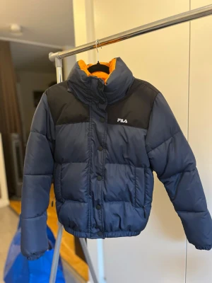 Mörkblå pufferjacka från FILA - Snygg mörkblå pufferjacka från FILA med svarta axelpartier och orange foder. Jackan har hög krage, dragkedja och knappar framtill samt två sidofickor med dragkedja. Perfekt för kalla vinterdagar och riktigt trendig look.