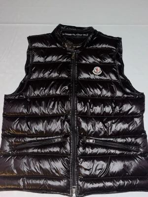 Svart glansig dunväst Moncler - Snygg svart dunväst från Moncler med glansigt material och klassisk logga på bröstet. Västen har hög krage, dragkedja framtill och två fickor med dragkedja. 
