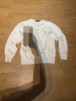 Vit stickad tröja Polo Ralph Lauren - Klassisk vit stickad tröja från Polo Ralph Lauren med rund halsringning och den ikoniska broderade loggan på bröstet. Tröjan har två hål vid var sin arm men annars är den fin 