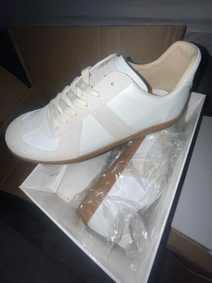 Maison margiela skor  - Stilrena vita sneakers med beige mockadetaljer och gummisula. Skon har klassisk låg profil, vita skosnören och en enkel design som passar till det mesta. Materialet är en mix av slätt läder och mocka för en snygg kontrast.